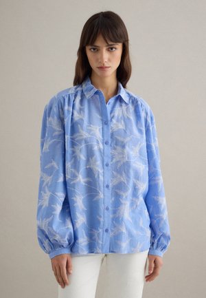 Frau trägt ein locker sitzendes blaues Hemd mit weißen Blattmustern und weiße Hose, steht vor einem schlichten hellen Hintergrund.