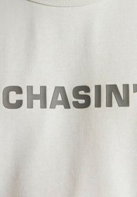 Sudadera gris claro con un audaz estampado de texto en gris oscuro que dice "CHASIN’", con un cuello redondo y textura suave.