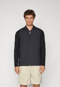 Black bomber-style jacket with a front zipper, long sleeves, and a smooth texture, paired with light beige shorts.