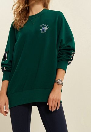 Femme portant un sweat-shirt oversize vert foncé avec un petit logo bleu et blanc sur la poitrine et des leggings noirs, montre-bracelet au poignet gauche.