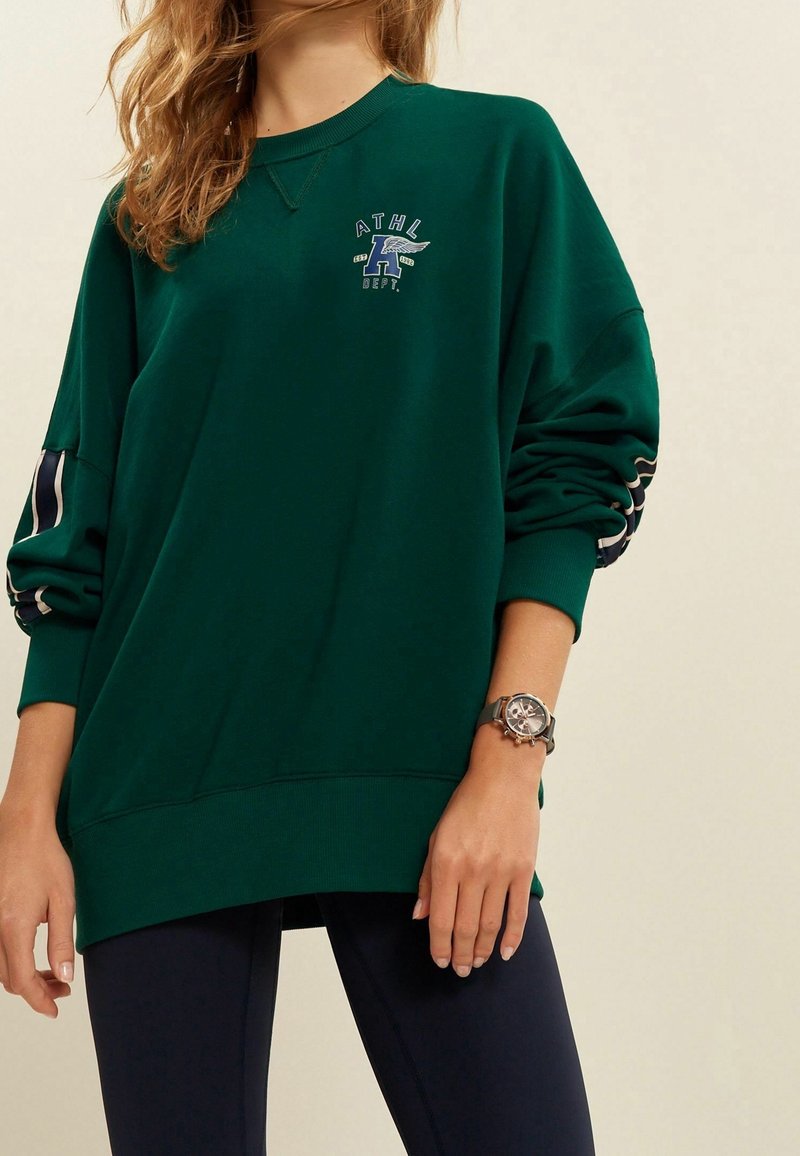 Femme portant un sweat-shirt oversize vert foncé avec un petit logo bleu et blanc sur la poitrine et des leggings noirs, montre-bracelet au poignet gauche.