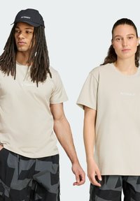 Camisetas Terrex beige con logo blanco en la parte frontal; el modelo a la izquierda lleva una gorra negra, ambos combinados con pantalones cortos oscuros con estampado.