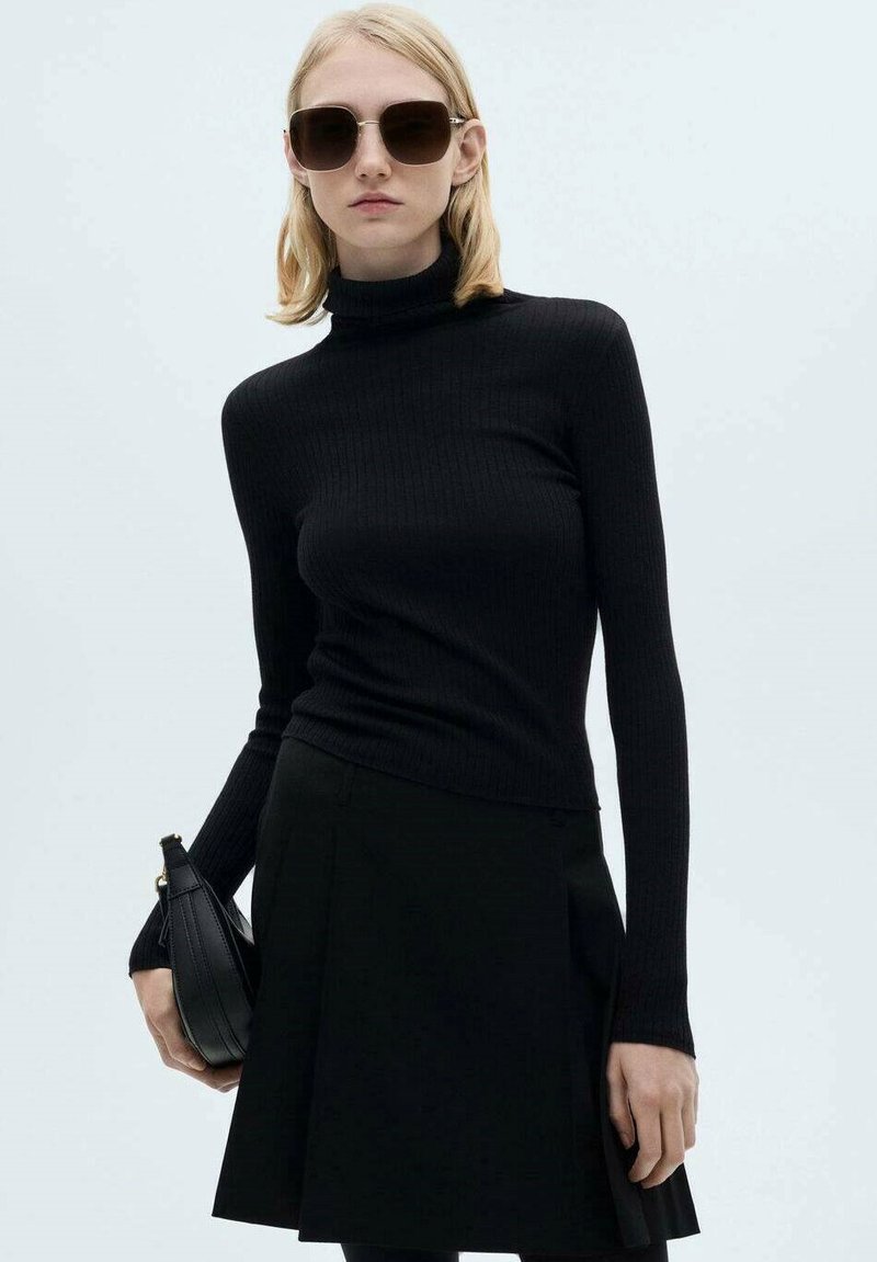 Pull à col roulé noir à côtes avec des manches longues, associé à une jupe noire, et tenant un sac à main noir. Les accessoires incluent des lunettes de soleil foncées.