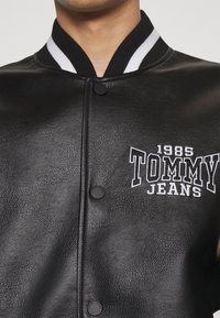 Musta nahkainen bomber-takki, jossa valkoraidallinen kaulus. Takissa on nappikiinnitys ja kirjailtu "1985 TOMMY JEANS" -logo rinnassa.
