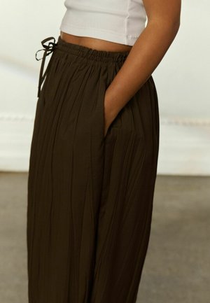 Persona che indossa pantaloni larghi marrone scuro con vita elasticizzata e coulisse, mano in tasca, abbinati a un top bianco corto.