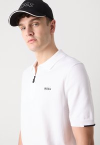 Hvit polo skjorte med glidelås og fint teksturert stoff. Har en grå "BOSS" logo og svarte detaljer på mansjettene og kragen.