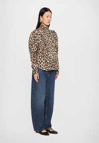 Blouse kuptues me print leopardi me detaje të thurura, e kombinuar me xhinse të errëta të lëshuara dhe këpucë flate të zeza. Veshjet theksohen me dërgues ari.