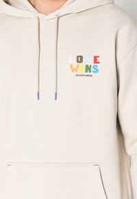 Felpa beige realizzata in tessuto morbido. Presenta la scritta "LOVE WINS" in lettere colorate. Include una tasca frontale e lacci con punte viola.
