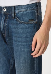 Jeans de denim azul oscuro con un corte clásico, que cuentan con cinco bolsillos, cierre de botón metálico y una etiqueta de G-Star Originals en la cintura.