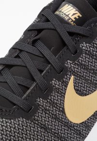 Zapatilla deportiva de Nike con un upper texturizado en negro y gris, cordones negros y un logo de swoosh dorado en el lateral. Presenta material sintético.