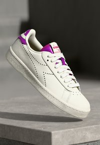 Hvit lær sneaker med teksturert overflate, lilla detaljer på hælen og tungen, samt perforerte detaljer langs sidene.