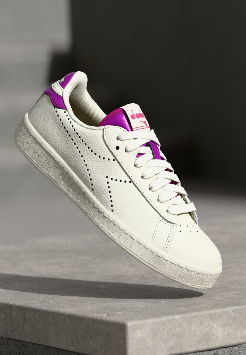Hvit lær sneaker med teksturert overflate, lilla detaljer på hælen og tungen, samt perforerte detaljer langs sidene.