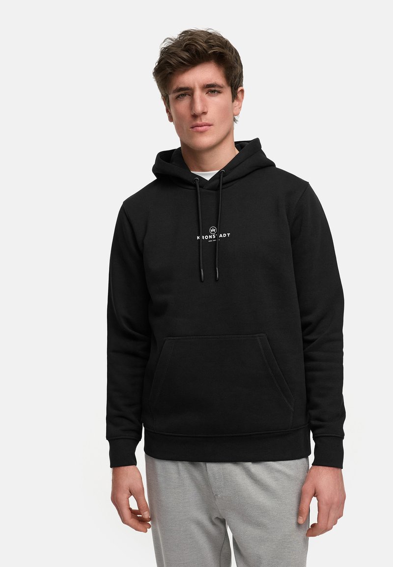 Svart hettegenser laget av mykt stoff, har en lomme foran, snøring på hetten og en hvit logo på brystet. Kombinert med grå joggebukser.