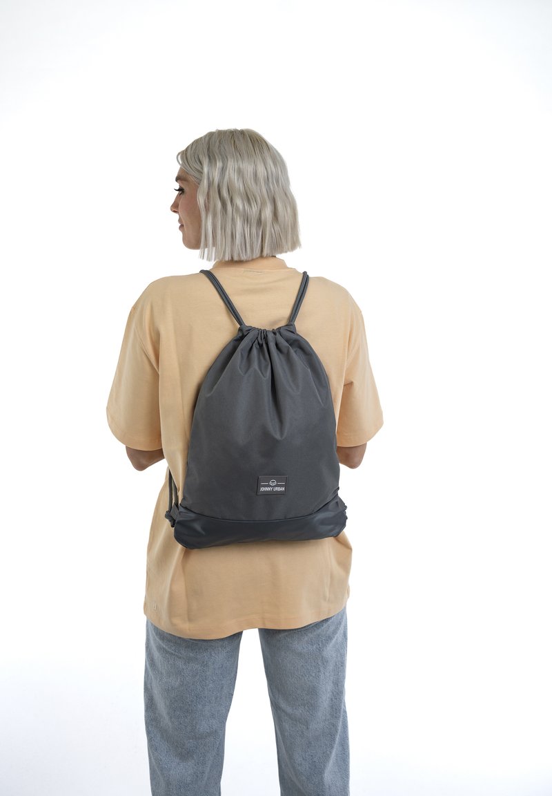 Mochila de cordón gris hecha de tela duradera, con parte superior ajustada, fondo plano y un parche con el logo en la parte frontal.