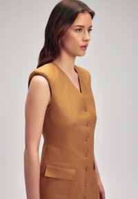 Vestido sin mangas de color mostaza con escote en V, cinco botones en el frente y un pequeño bolsillo lateral. Material de lino texturizado.