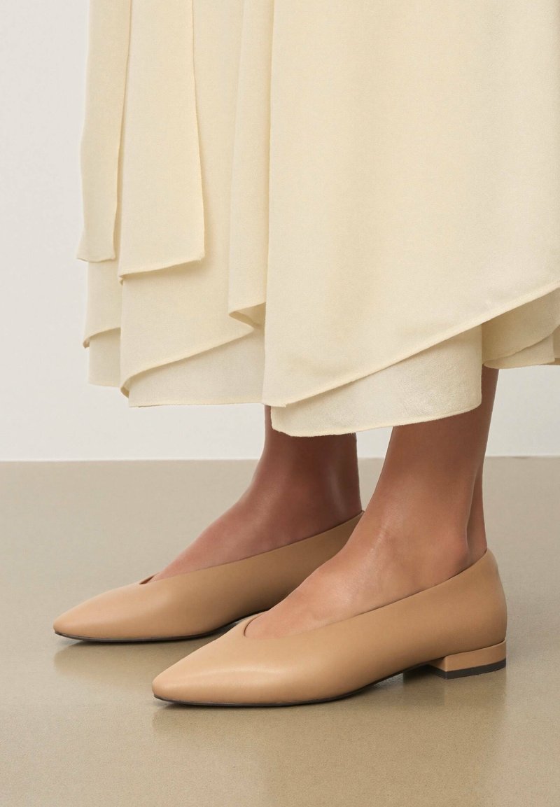Ballerines en cuir nude avec bout pointu et dessus ouvert, dotées d'un petit talon bloc. Jupe crème longue jusqu'au sol partiellement visible au-dessus des chaussures.