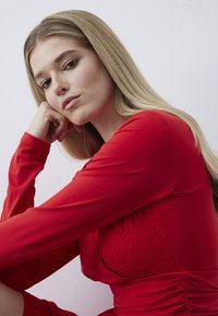 Robe rouge à manches longues avec un corsage en dentelle texturée, silhouette ajustée et détail froncé à la taille, sur fond blanc.