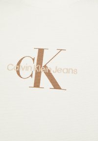 Logo di Calvin Klein Jeans con grandi lettere marroni "CK" e testo beige più piccolo su uno sfondo di tessuto bianco texturizzato.