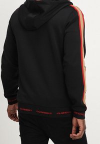 Muž v černé mikině s kapucí s červenými a béžovými pruhy na rukávech, nápisem "GLORIOUS" na manžetách a lemu, stojící zády ke kameře.