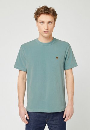 Giovane uomo con capelli corti castano chiaro che indossa una t-shirt a maniche corte a coste color turchese con un piccolo logo a forma di albero marrone sul petto.