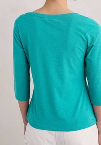 Teal topp med trekvartsärmar och rund halsringning, tillverkad av mjukt tyg, slät textur och en åtsittande silhuett.