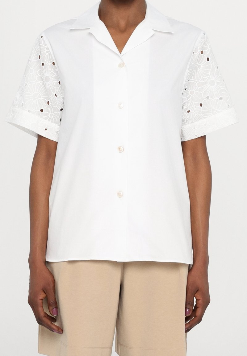 Blouse blanche à manches courtes avec manches en dentelle florale et boutons à l'avant, portée avec un short beige sur une personne à la peau foncée.