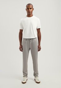 Grijze joggers met een drawstring taille, gecombineerd met een eenvoudig wit t-shirt. Het model staat tegen een neutrale achtergrond en draagt witte sneakers.