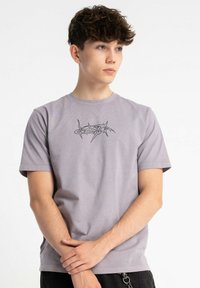 Jasnofioletowa bawełniana koszulka, z okrągłym dekoltem, krótkimi rękawami, z czarnym abstrakcyjnym wzorem na przodzie. Gładka tekstura, luźny krój.