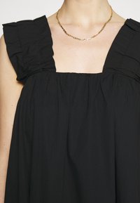 Černé šaty s čtvercovým výstřihem a volánovými rameny. Vyrobené z lehkého materiálu s plisovanou texturou a volným střihem. Součástí je zlatý řetízkový náhrdelník.