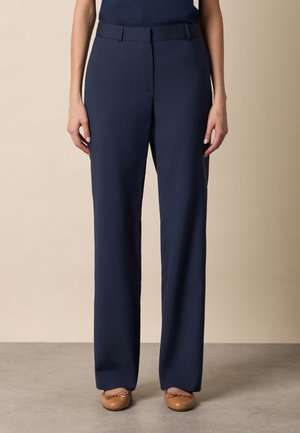 Marineblauwe, op maat gemaakte, recht gesneden broek gedragen met beige ballerina's, getoond op een persoon die tegen een effen achtergrond staat.