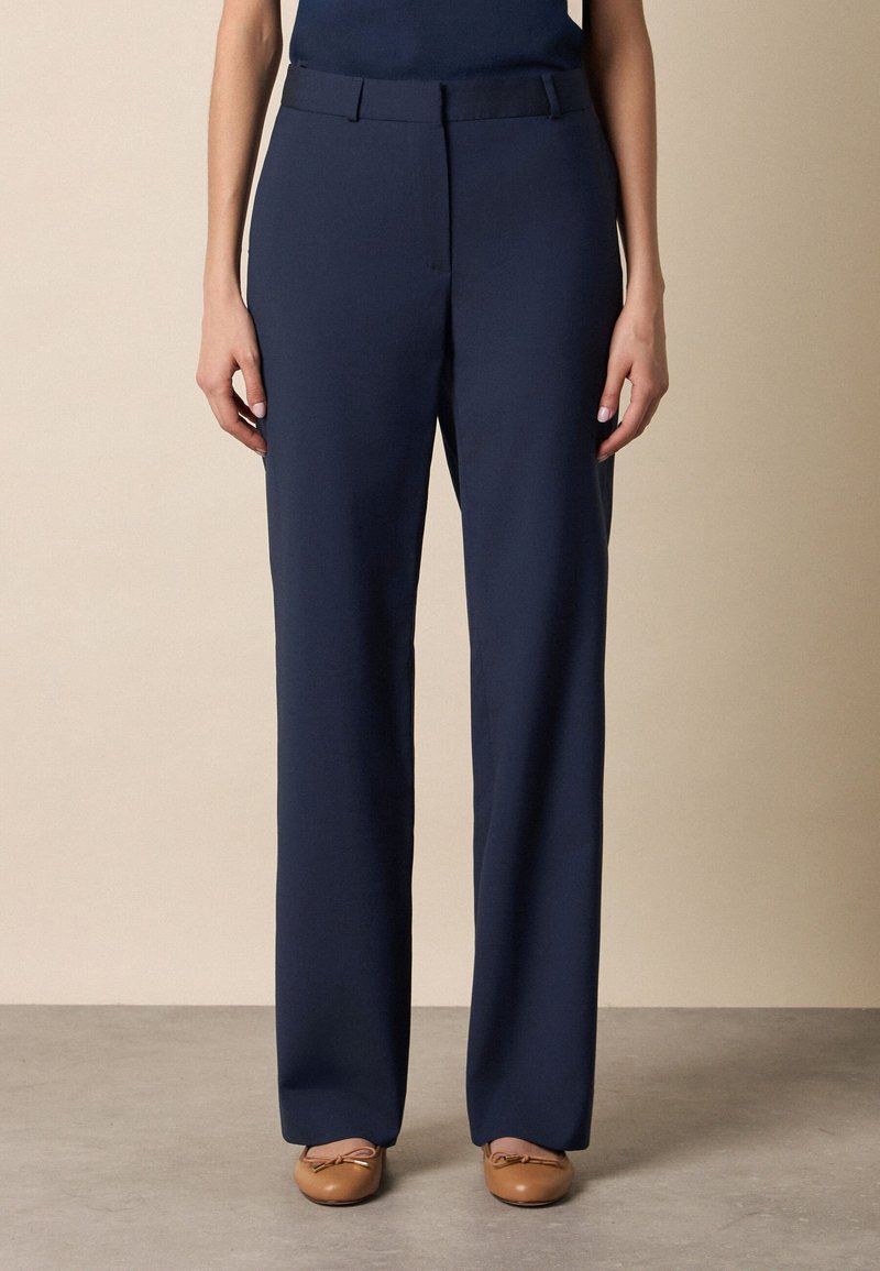 Marineblauwe, op maat gemaakte, recht gesneden broek gedragen met beige ballerina's, getoond op een persoon die tegen een effen achtergrond staat.