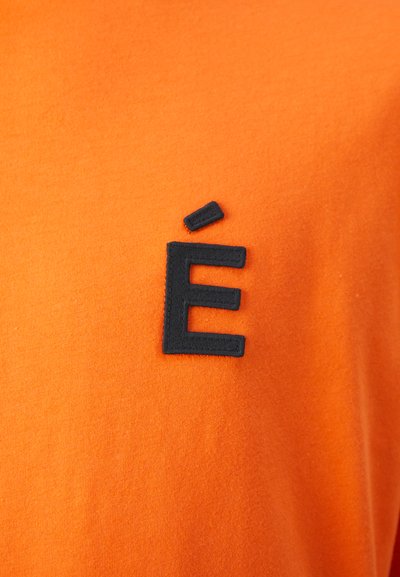 Orange cotton shirt featuring a black embroidered accent with the letter "É" on the left side. Soft texture with a smooth finish.