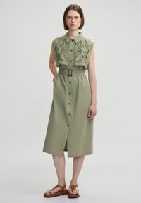 Abito midi verde oliva con top ricamato corto, vita con cintura e apertura frontale con bottoni. Presenta tasche laterali e uno spacco fino al ginocchio.