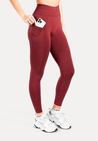 Bordeauxrode leggings met hoge taille en een zijzak. Gemaakt van rekbare stof, met een gladde textuur en een aansluitende pasvorm. Draagt bij witte sneakers.