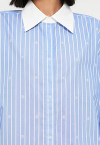 Svetlomodrá košeľa s bielymi vertikálnymi pruhmi a bielym golierom. Na látke je jemný opakujúci sa vzor loga.