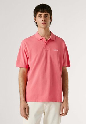 Joven con una camisa polo de manga corta color coral liso y pantalones beige claros, de pie frente a un fondo blanco.