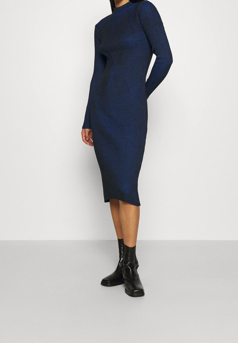 Marinblå ribbad klänning med långa ärmar, figurnära silhuett och sidofickor, matchad med svarta ankelstövlar. Slät struktur med vertikala ränder.