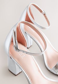 Paire de sandales argentées à talons hauts avec brides à la cheville et talons carrés épais sur un fond clair.