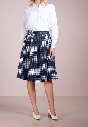 Mujer vestida con una camisa blanca de botones, falda plisada azul marino con un patrón a cuadros blancos y tacones blancos, de pie sobre un suelo neutro.
