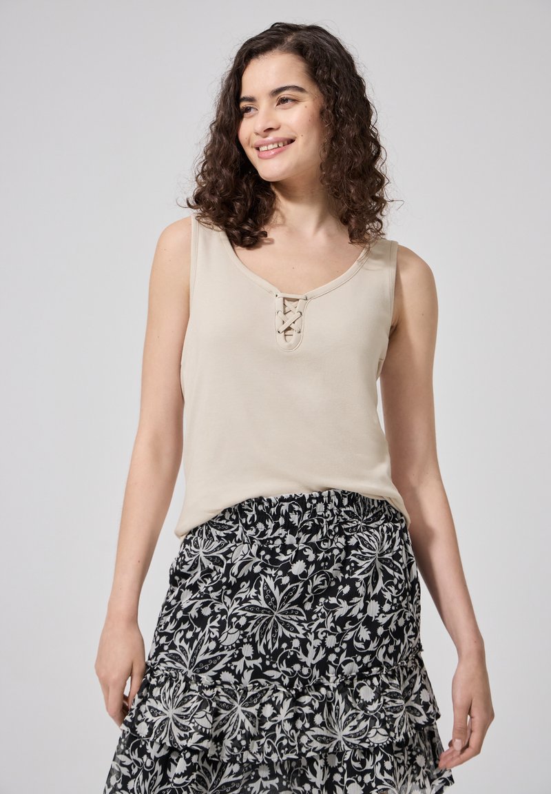 Femme aux cheveux bouclés portant un haut beige sans manches avec un détail à lacets et une jupe à motif floral noir et blanc, souriante et regardant sur le côté.