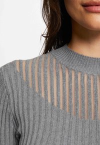 Grauer Pullover mit gerippter Textur, der transparente Einsätze mit vertikalen Streifen im oberen Bereich und einen runden Ausschnitt aufweist.