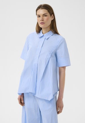 Femme portant une chemise ample bleu clair à manches courtes et un pantalon large assorti avec des rayures blanches verticales, debout devant un fond uni.