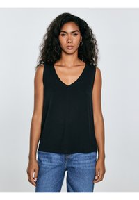 Jonge vrouw met lang, krullend haar, gekleed in een zwart mouwloos topje met V-hals en blauwe spijkerbroek, staand tegen een effen lichte achtergrond.