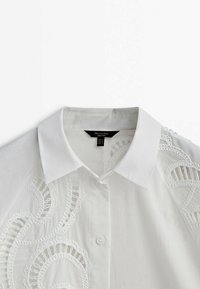 Chemise blanche boutonnée avec un col classique, présentant des détails en dentelle élaborée sur un côté et un tissu doux et texturé.