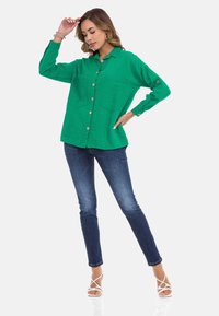Groene button-up shirt met twee borstzakken, lange mouwen en een relaxed fit, gecombineerd met donkerblauwe skinny jeans en witte sandaal.