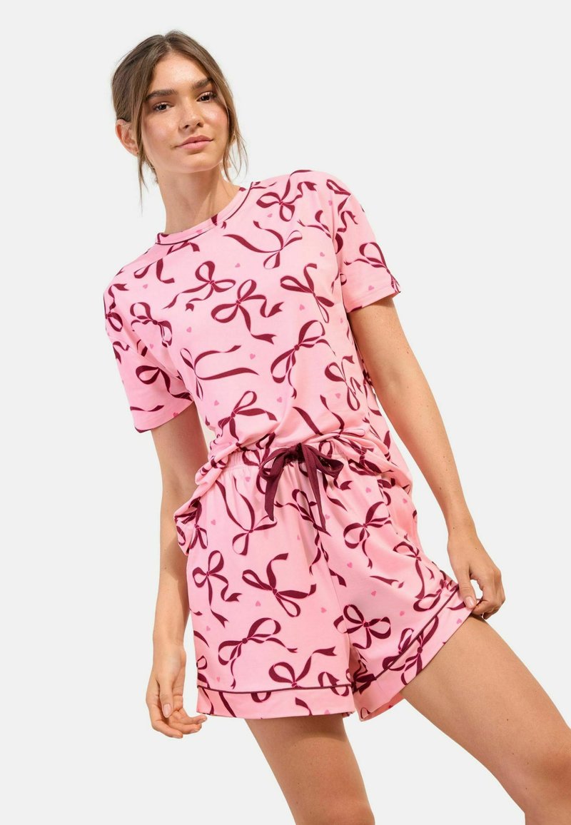 Nuori nainen pukeutuneena vaaleanpunaiseen lyhythihaiseen puseroon ja shortseihin, joissa on tummanpunaiset rusetti- ja sydänkuviot, seisoo yksinkertaisen vaalean taustan edessä.