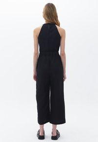 Svart jumpsuit med halterneck, vida ben och plisserad midja. Slät tyg med en subtil glans, med en nyckelhål detalj bak.