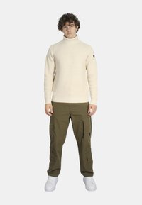 Maglione a collo alto a coste crema abbinato a pantaloni cargo oliva con multiple tasche e cerniere, indossati con scarpe da ginnastica bianche.