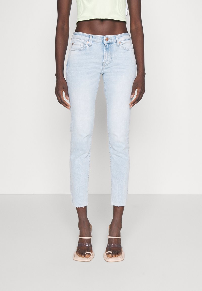 Hellblaue, figurbetonte Jeans mit fransigem Saum, fünf Taschen und Knopfverschluss, kombiniert mit klaren Sandalen mit Absatz.