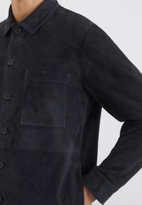 Schwarze Wildlederjacke mit Kragen, Knopfverschluss und einer einzigen Brusttasche. Sie zeichnet sich durch eine glatte Textur und eine taillierte Passform aus.